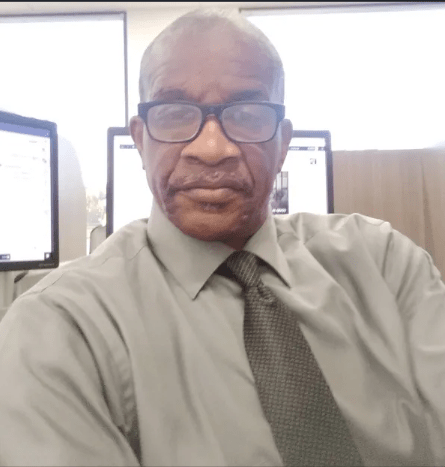 A man in a gray dress shirt and tie poses for a selfie at a workstation with computer monitors in the background